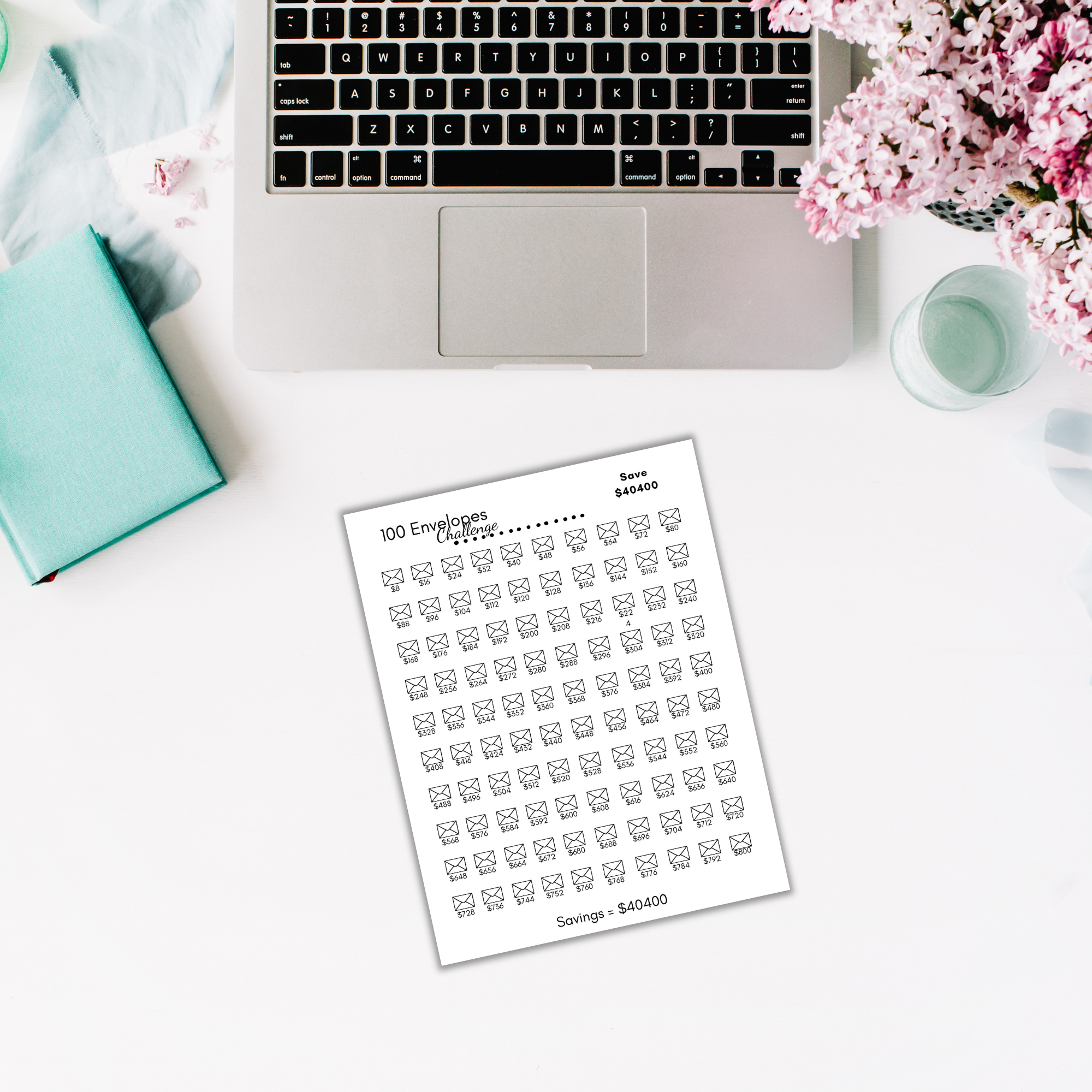 An open laptop sits on a white desk next to flowers, a teal notebook, and The 100 Envelope Challenge tracker templates for the savings plan.
