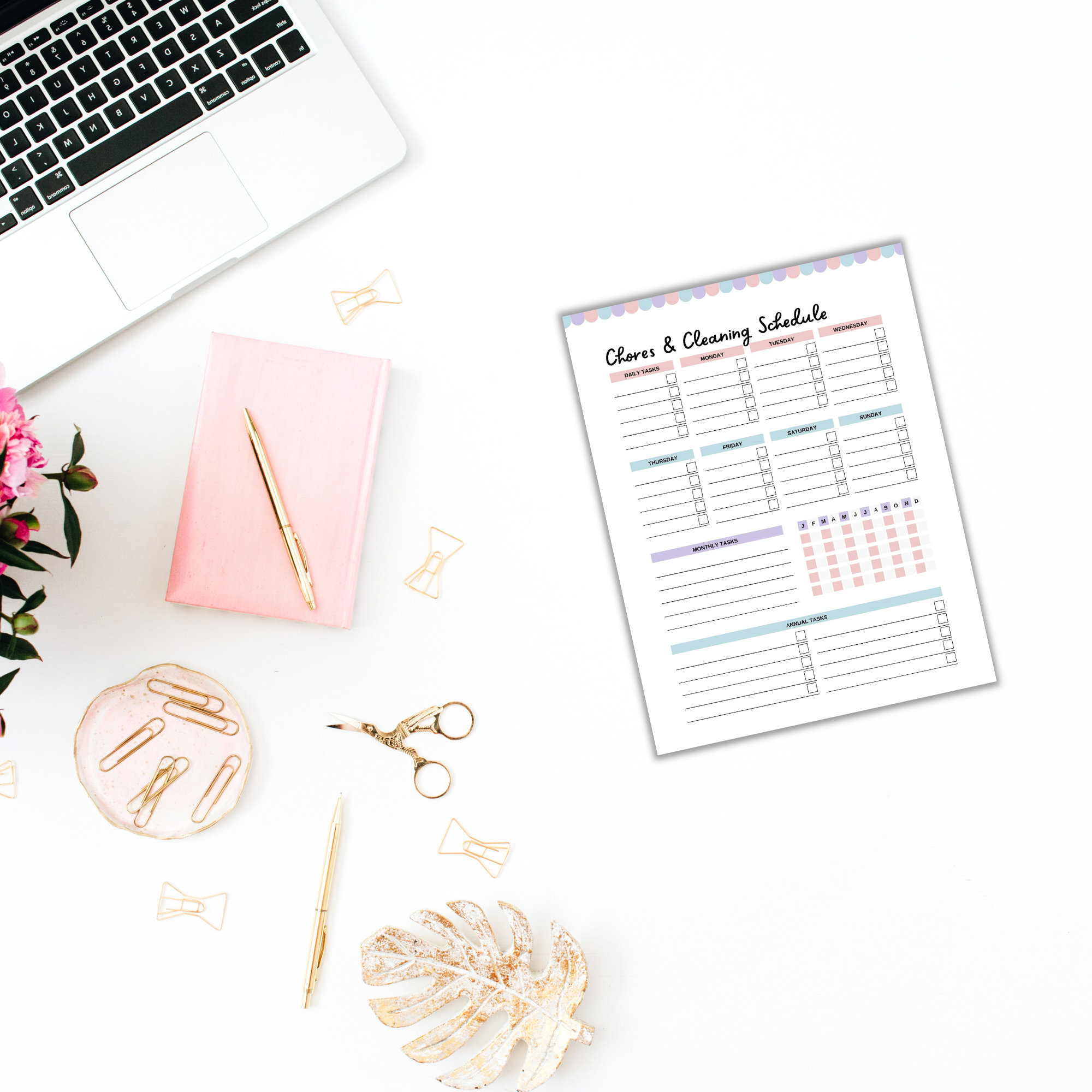 A cozy desk setup includes a laptop, The Cottagecore Cleaning Planner in pink, gold paper clips, scissors, and a pen. To add a touch of cottagecore charm, there's leaf decor and the planner neatly displaying chores and cleaning schedules on paper.