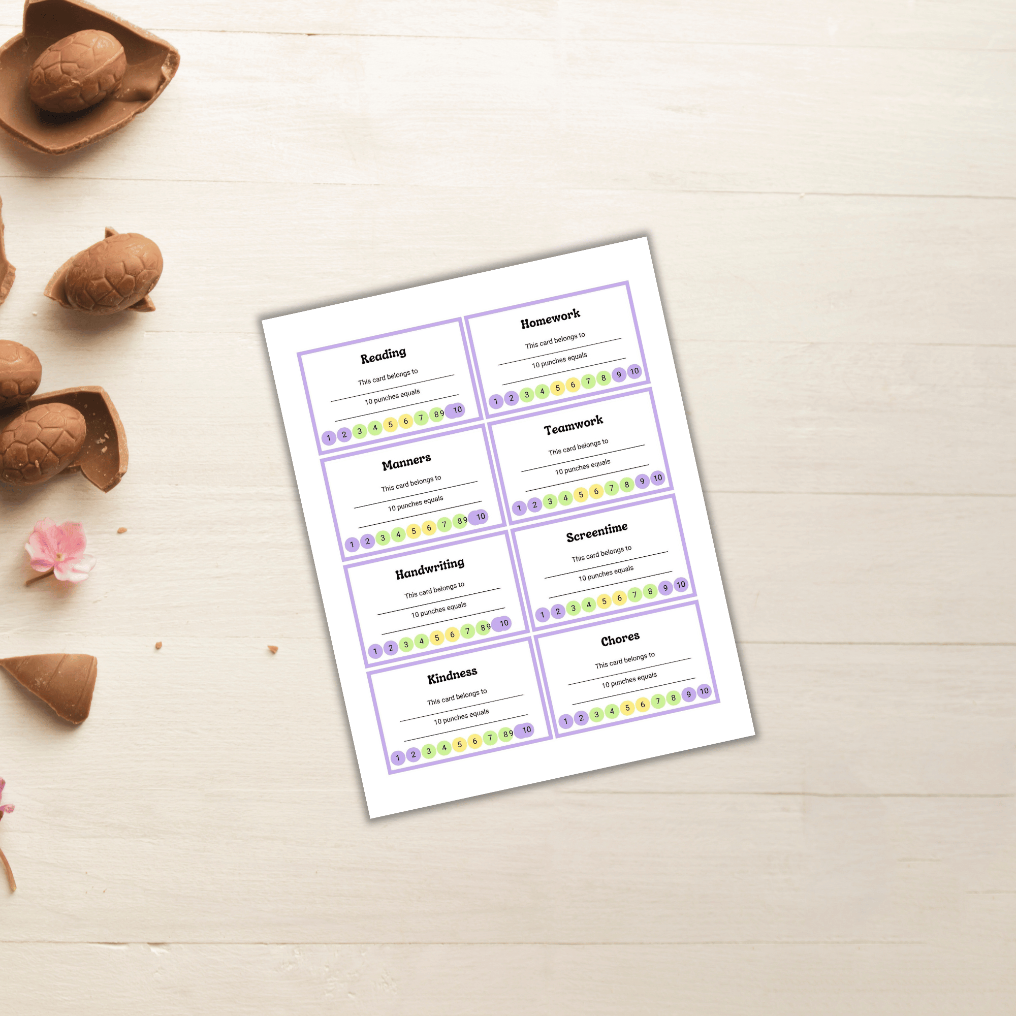 Eight purple and white chore cards from the Free PLR: Spring Refresh Bundle are placed on a light wooden surface, surrounded by chocolate egg halves and pink flowers. The cards include tasks such as reading, homework, manners, and chores.